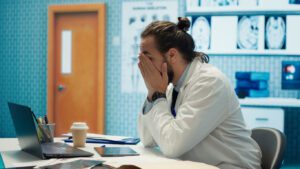 Tired worried doctor working after hours to find the diagnostic for a patient, feeling very stressed and trying to examine x ray results in medical cabinet. Frustrated overworked medic. 3 LEADING CAUSES OF PRACTICE OWNER BURNOUT… AND HOW TO SOLVE IT